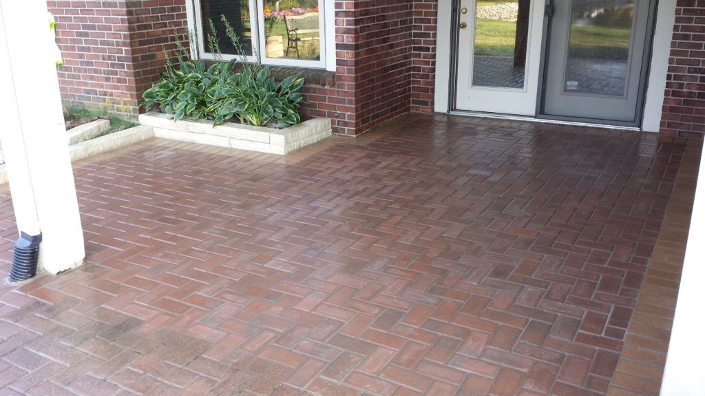 A red brick patio with a herringbone pattern leads to a building entrance with double doors and a brick planter.
