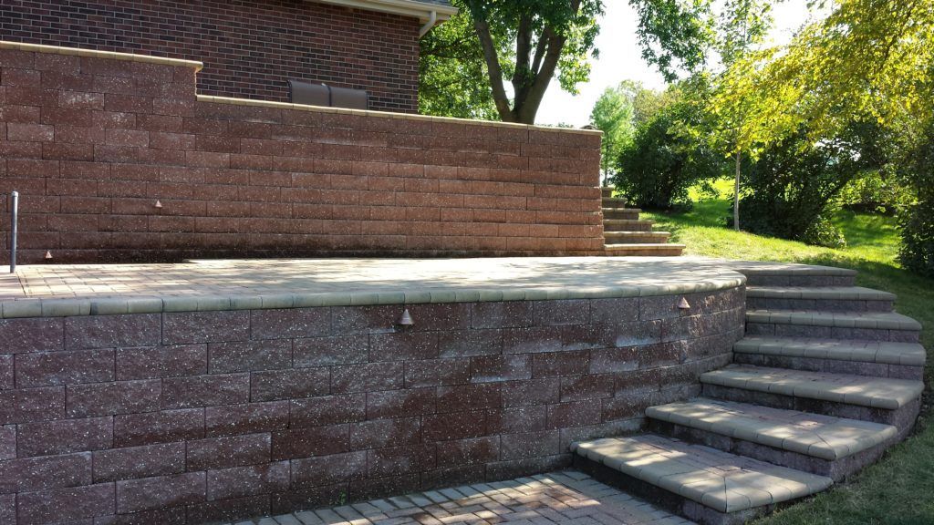 A stone patio with a retaining wall and curved steps, located outdoors next to a brick house.