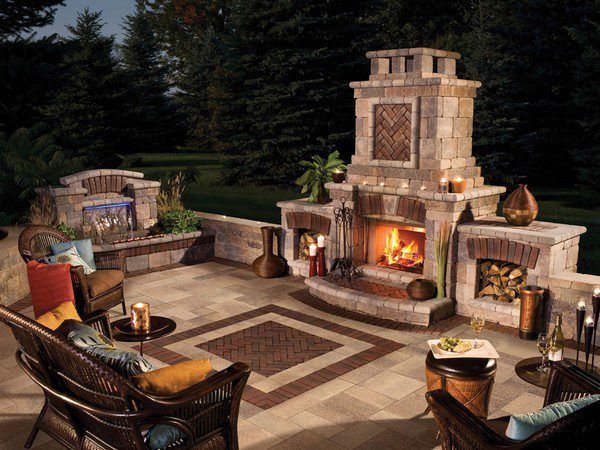 An outdoor patio at twilight featuring a stone fireplace, integrated seating, and wicker furniture with colorful cushions.
