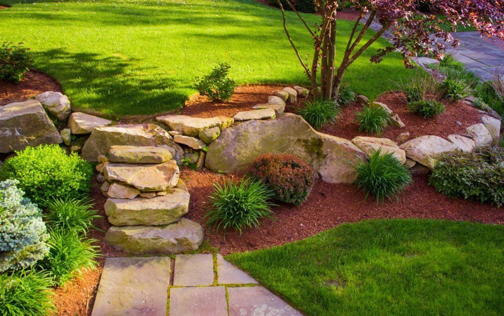 A stone stairway transitions between two tiers of a grassy lawn, accented by boulders and red mulch landscaping.