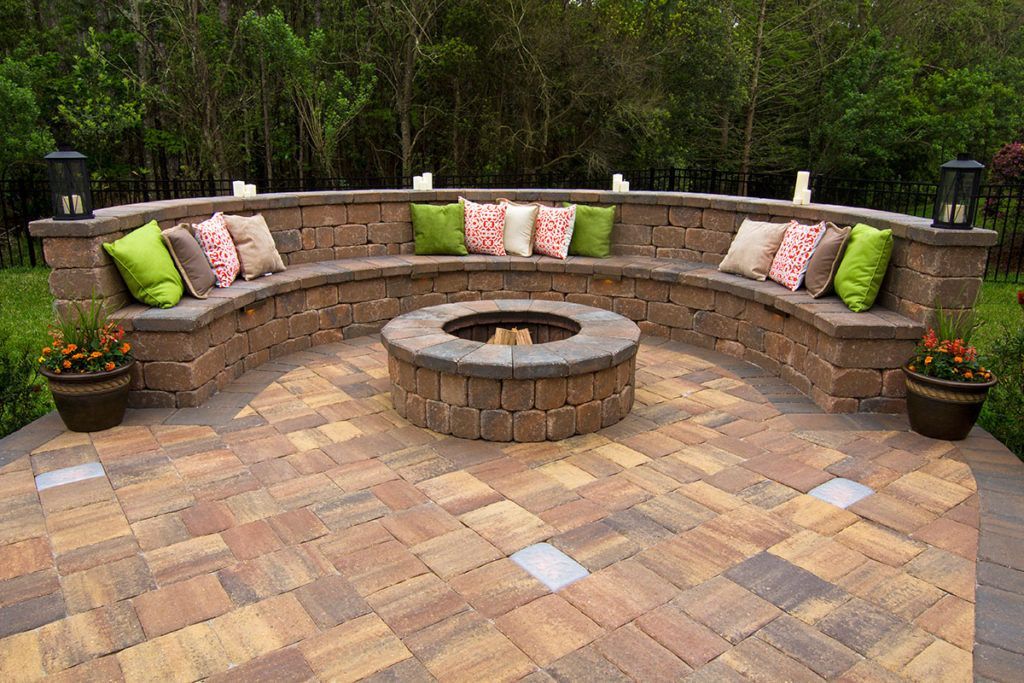 A stone patio features a semi-circular built-in seating wall with colorful throw pillows surrounding a round fire pit.