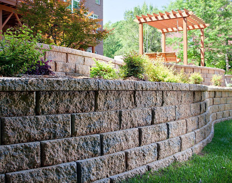 A multi-tiered stone retaining wall curves through a landscaped backyard with a wooden pergola in the background.