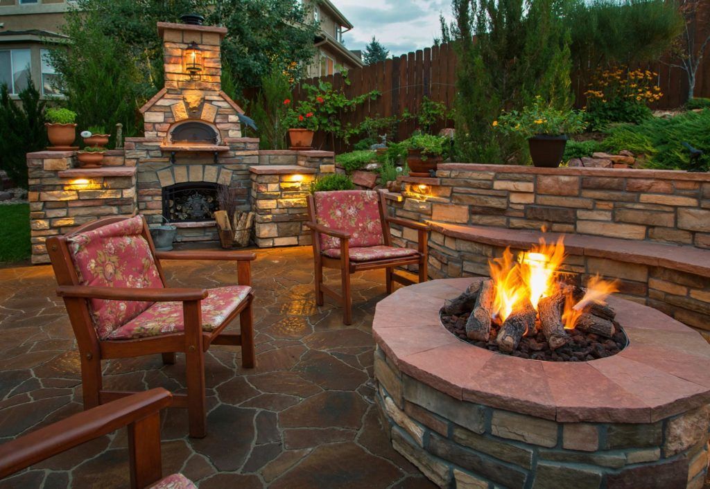 A stone fire pit burns on a backyard patio with outdoor chairs, a stone fireplace, and a curved seating wall at dusk.