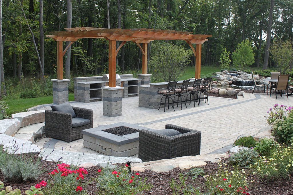 A stone patio with an outdoor kitchen, pergola, fire pit, and cushioned seating set against a wooded backdrop.