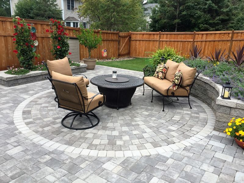 A circular stone patio with a fire pit, two armchairs, and a loveseat, set in a backyard with a wooden fence and plants.