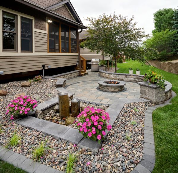 A backyard patio features a circular fire pit, stone fountain, gravel landscaping, and vibrant pink flowers by a house.