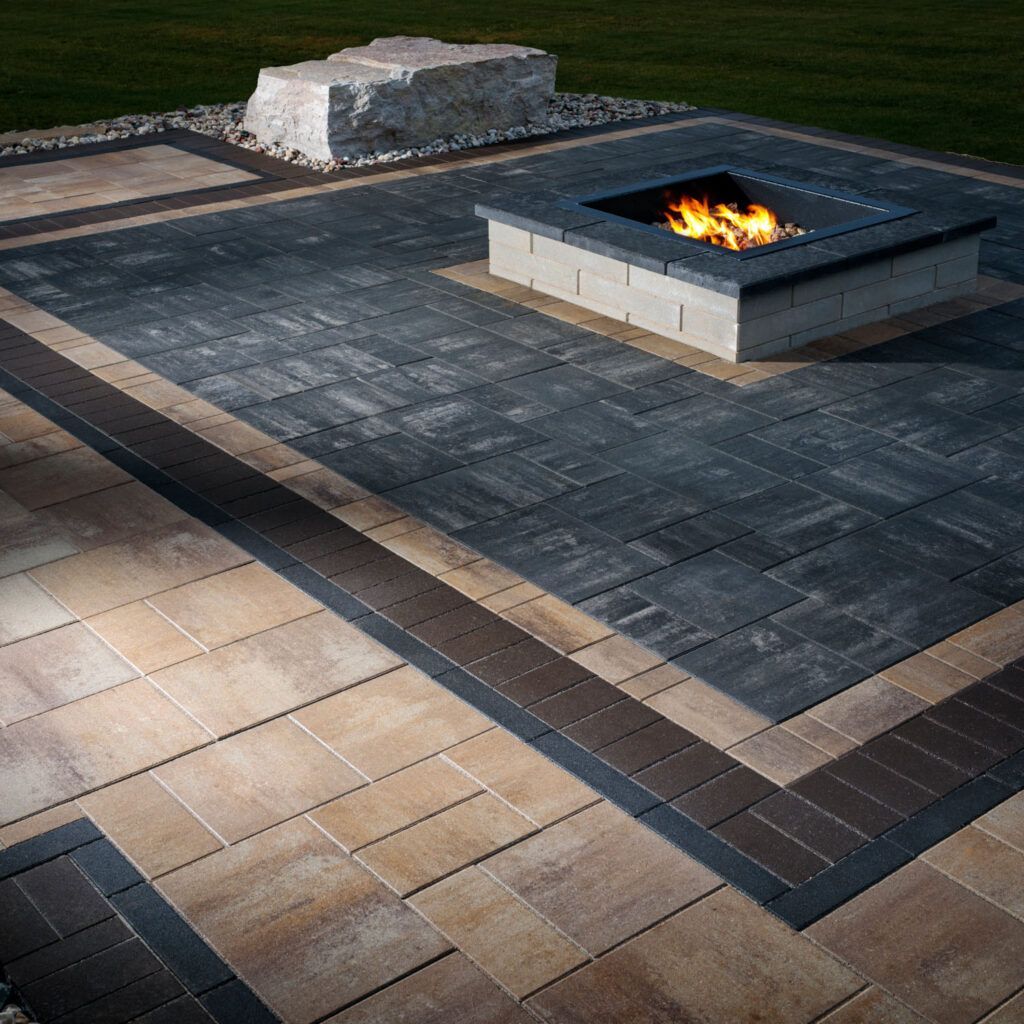 A square fire pit burns on a patio of dark and tan rectangular pavers, featuring a large stone accent in the background.