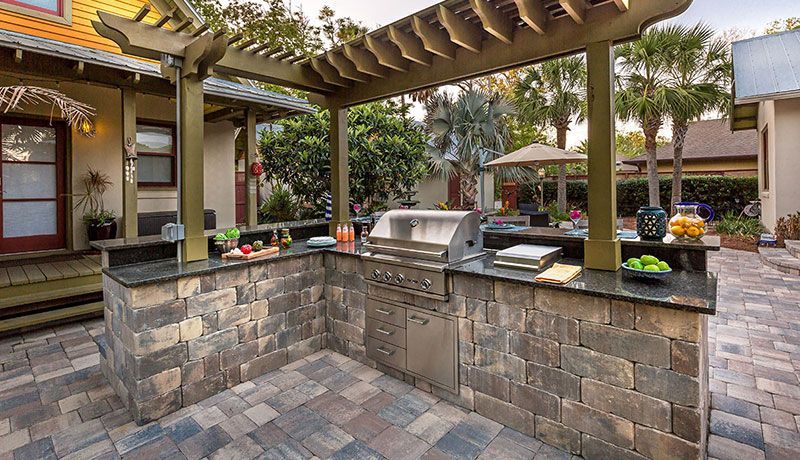 Outdoor kitchen with stone counters, a stainless grill, and a wooden pergola on a paved patio next to a house.