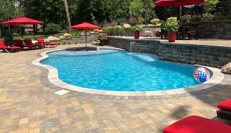 A backyard pool with bright blue water, paved patio, red umbrellas, and a small US-flag patterned beach ball floating.