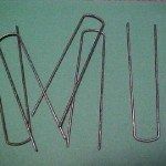 Three U-shaped metal landscape staples arranged on a plain, solid green background.
