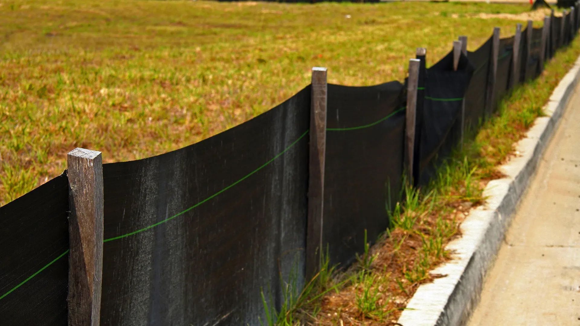 Black silt fence lining a grassy roadside beside a concrete curb and pavement