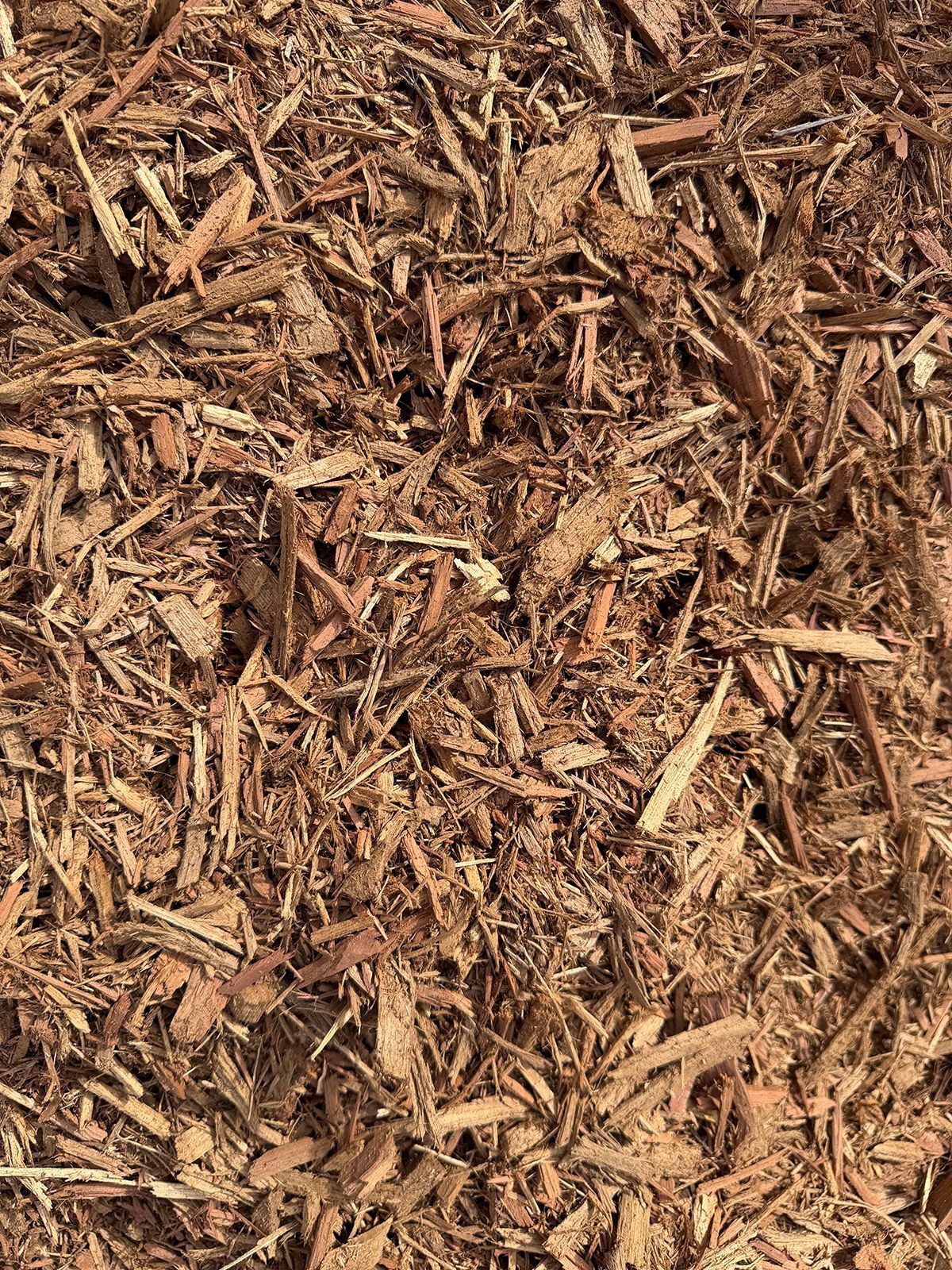A close-up view of brown wood mulch, consisting of coarse, shredded bark and wood chips covering the ground.