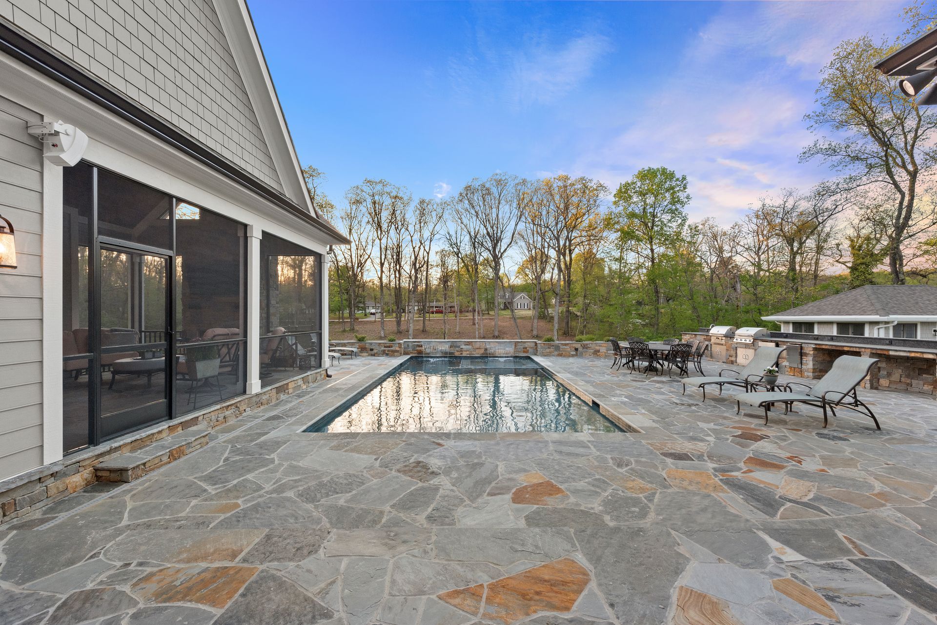 A luxury patio features a rectangular swimming pool, stone tile flooring, a screened porch, and lounge chairs at dusk.