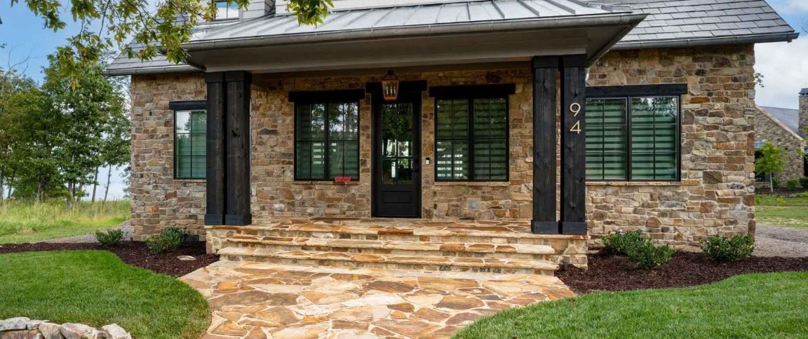 A stone house with a covered porch, dark window frames, and a stone walkway in a grassy, rural setting.