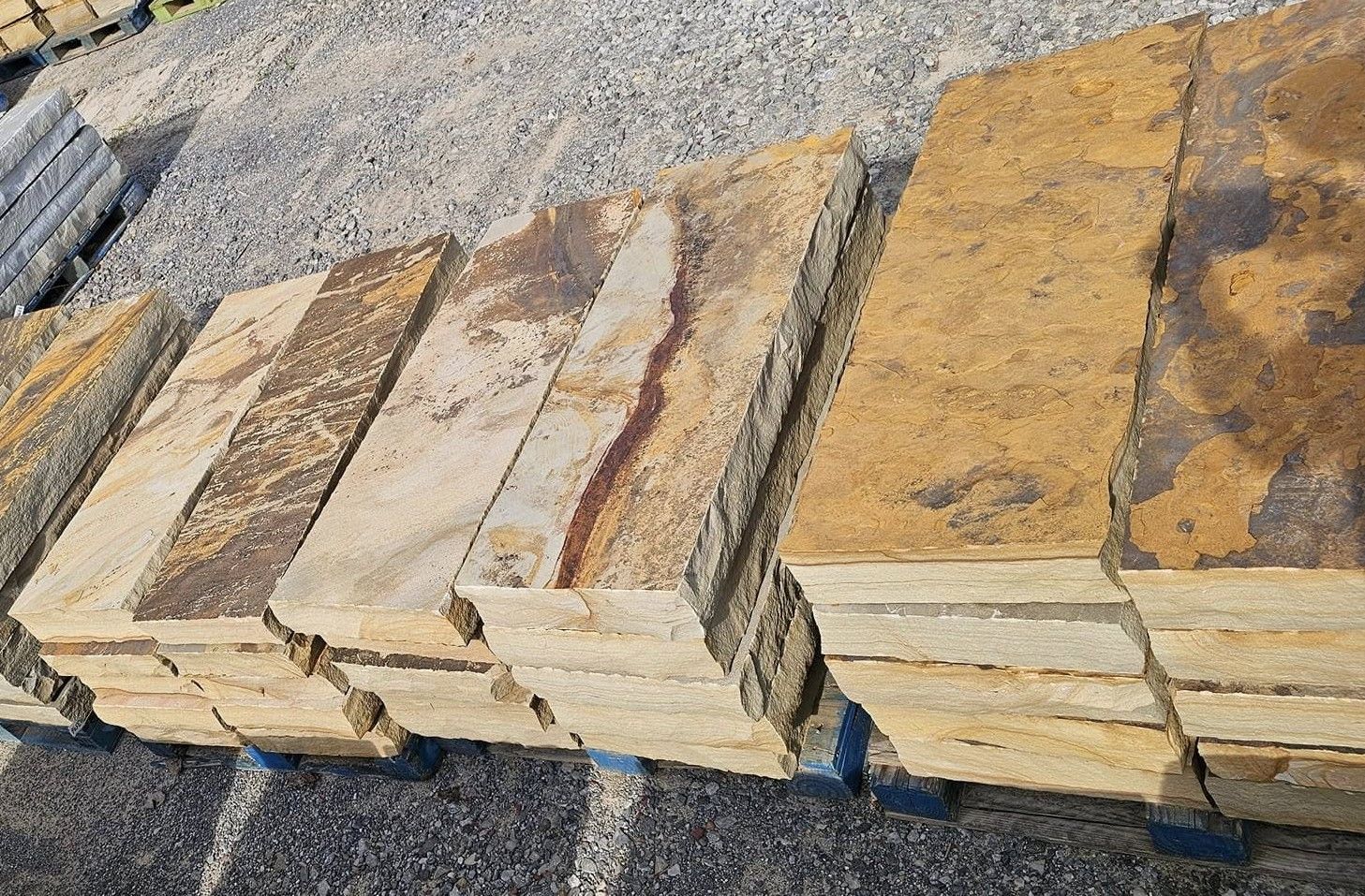 A stack of rectangular, natural-toned stone slabs with textured edges arranged on pallets on a gravel surface.