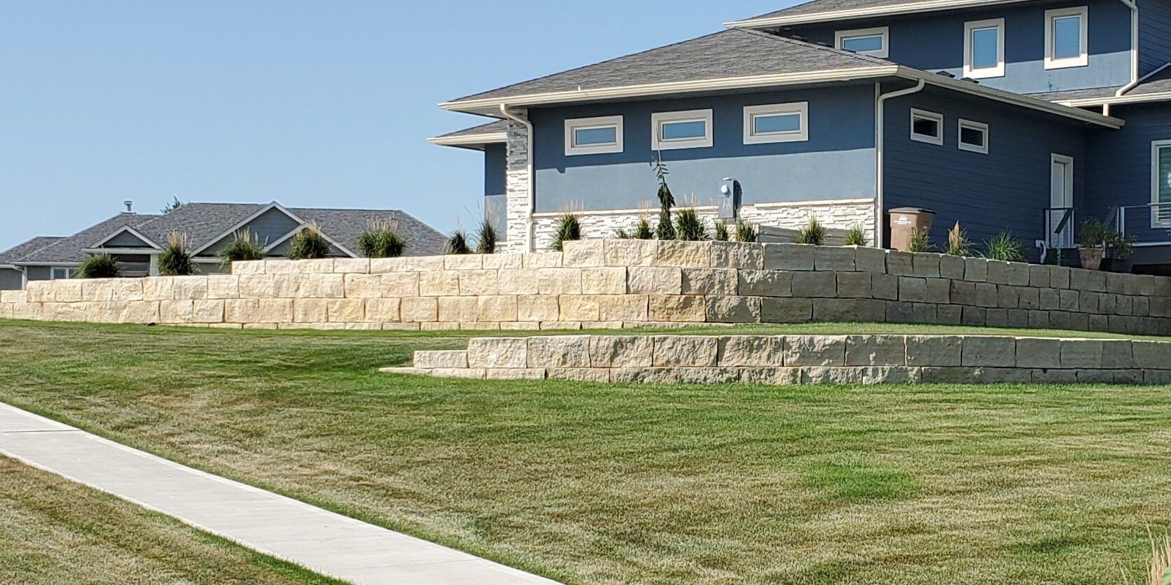 A modern blue house sits behind a tiered, light-colored stone retaining wall in a suburban grassy yard.
