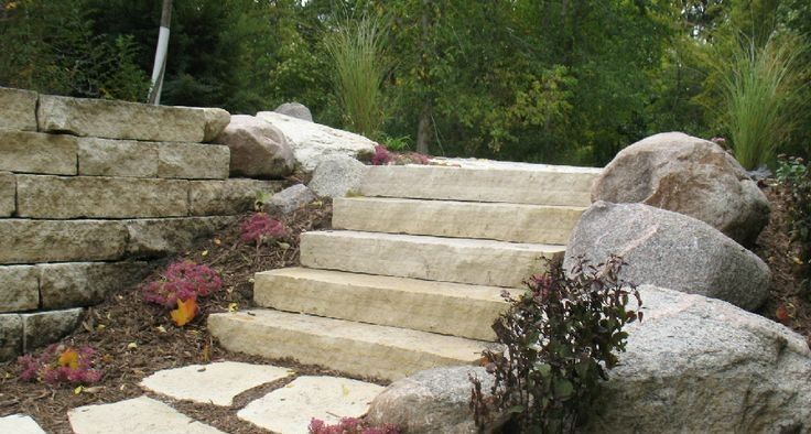 Stone steps lead up a gentle slope, flanked by a stacked stone retaining wall, large decorative boulders, and greenery.
