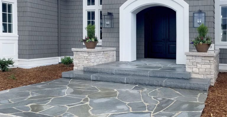 A stone entryway with steps leading to a dark front door, flanked by potted plants on stone pillars and gray shingle siding.