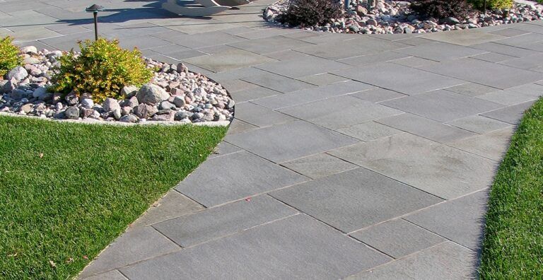 A curved stone walkway made of large gray pavers, flanked by green grass and landscaping beds with rocks and bushes.