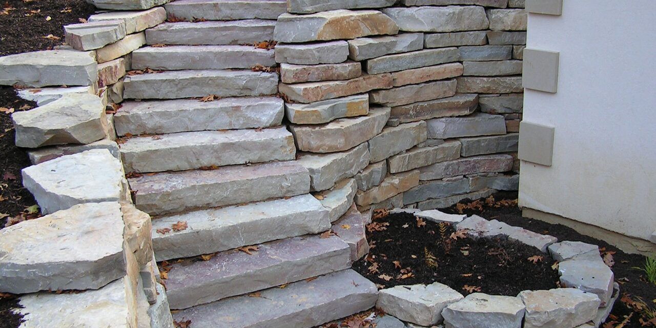 Curving stone steps lead upward alongside a matching stacked stone retaining wall next to a white building exterior.