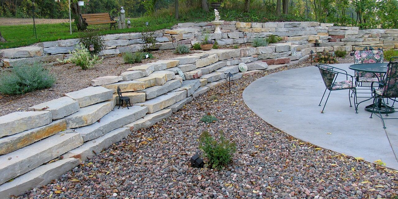 A tiered stone retaining wall separates a gravel garden landscape from a circular concrete patio with outdoor furniture.
