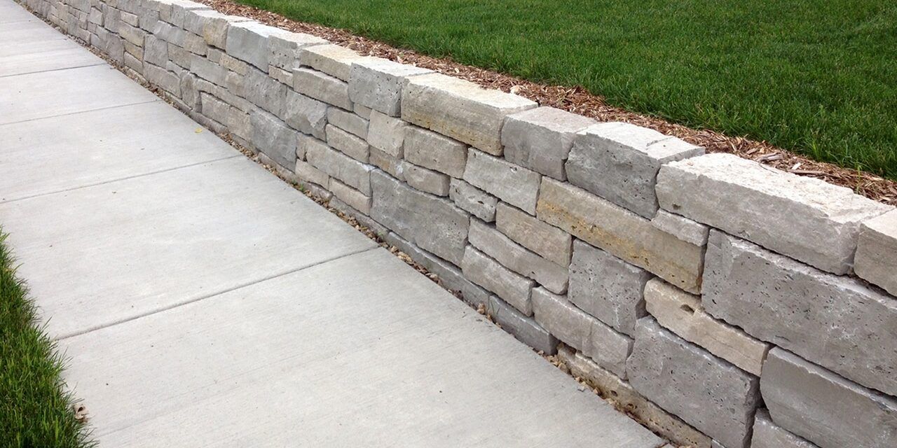 A low, gray stone retaining wall separates a concrete sidewalk from a patch of green grass.