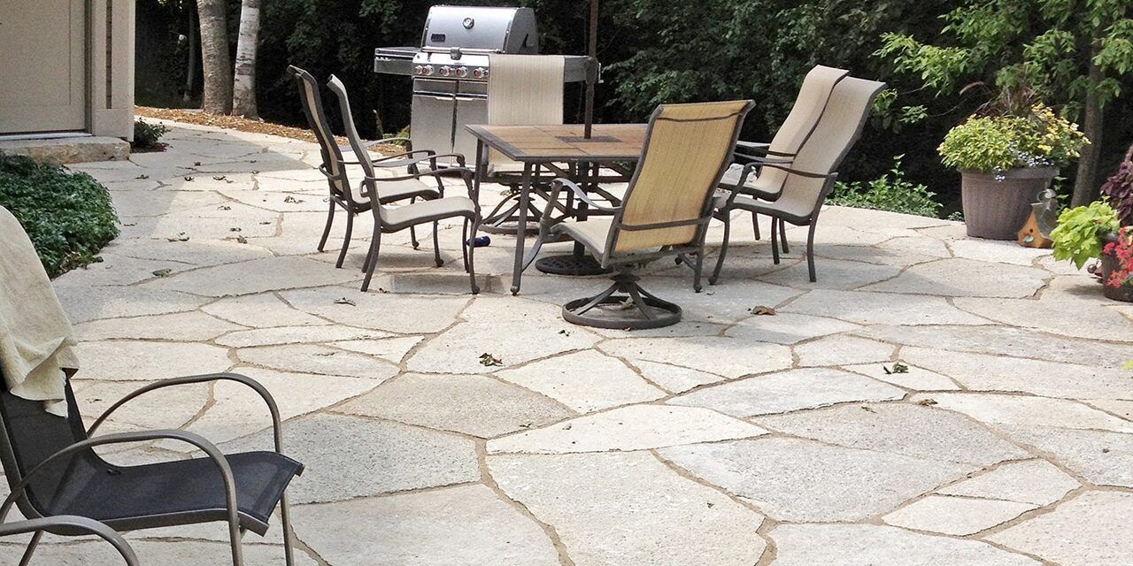 A stone patio featuring a dining set with chairs, a gas grill in the background, and potted greenery.