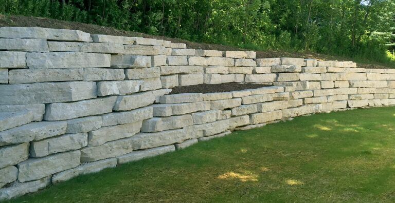 A light gray, dry-stacked stone retaining wall sits at the edge of a green lawn in front of a tree line.