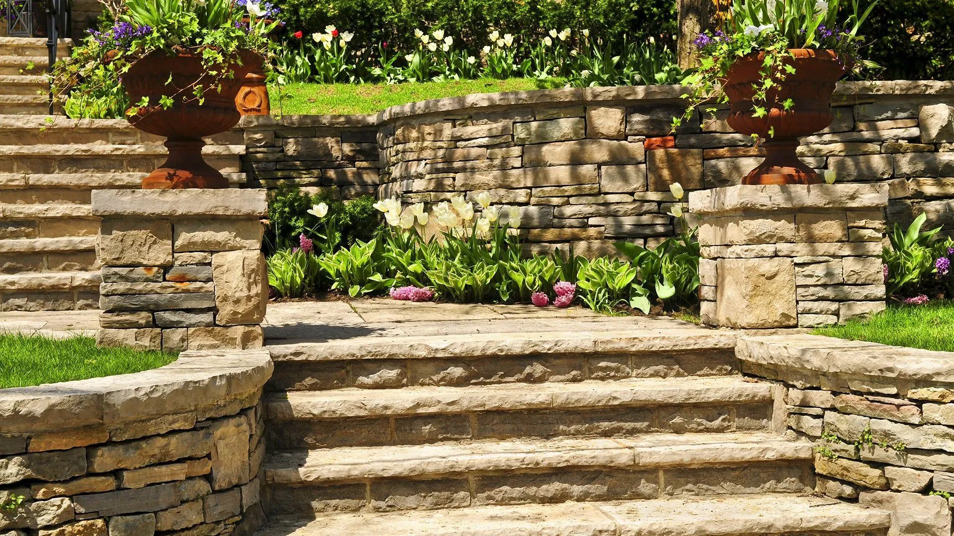 Stone garden steps leading to a raised terrace with flowering plants and potted urns on brick pillars