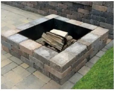 A square backyard fire pit made of stacked gray and tan stone blocks with a black metal liner and logs inside.