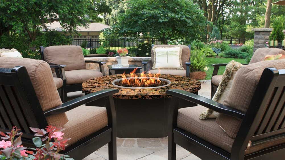 A circular fire pit with a lit flame surrounded by four cushioned armchairs on a paved patio in a lush backyard.