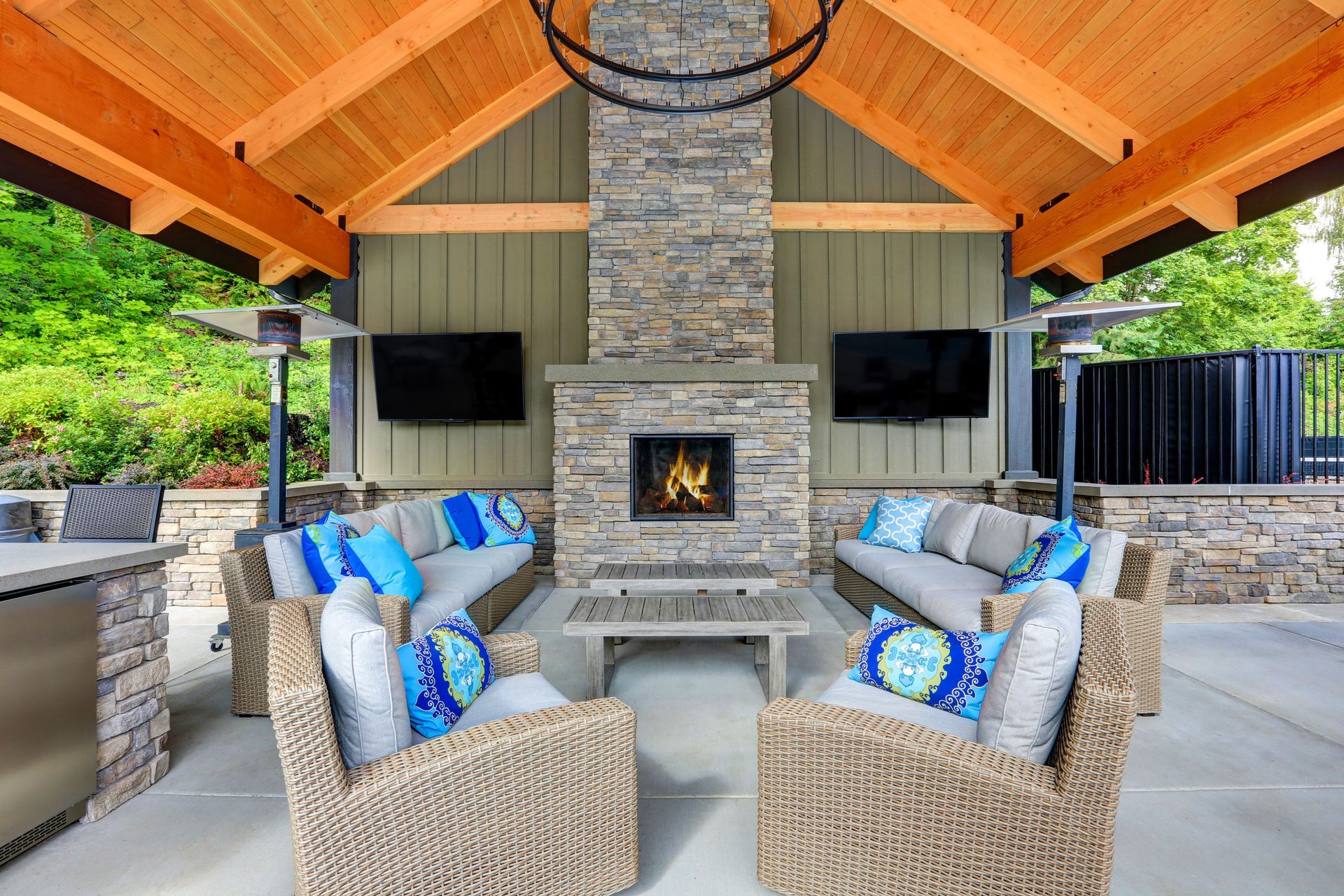 Covered outdoor living room with stone fireplace, wicker seating, and blue-gray cushions under a wooden roof