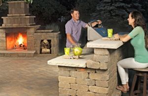 A person stands behind a stone patio bar while another person sits on a stool, with a glowing outdoor fireplace nearby.