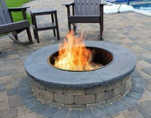 A circular stone fire pit with a bright flame, surrounded by dark chairs on a paved patio.