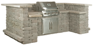 An L-shaped outdoor kitchen island made of gray stacked stone with a built-in stainless steel grill and cabinet doors.