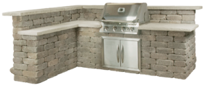 An L-shaped outdoor kitchen island made of gray stacked stone with a built-in stainless steel grill and cabinet doors.