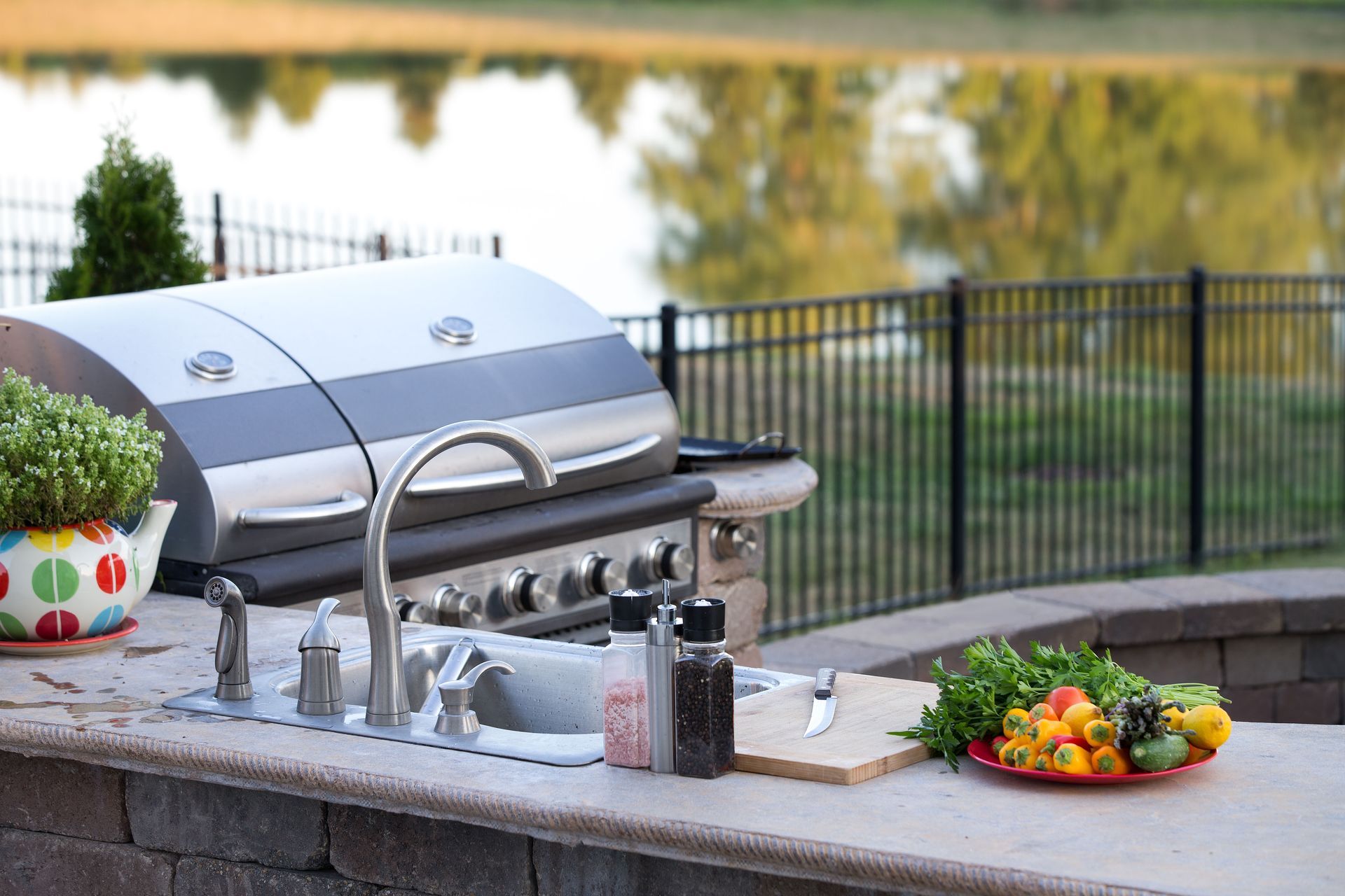 Outdoor patio with a stainless steel grill, sink, condiment bottles, and a bowl of food by a poolside fence