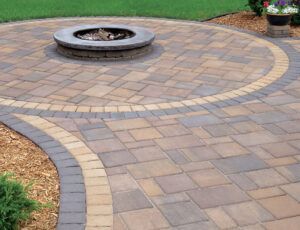 A circular fire pit centered on a multi-toned patio made of tan and brown pavers with a curved decorative border.