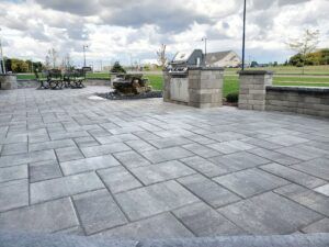 A gray paver patio with a built-in stone grill station and outdoor seating area under a cloudy sky.