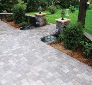 A stone patio with a gray paver pattern, featuring two small fountain basins built into stone columns in a garden setting.