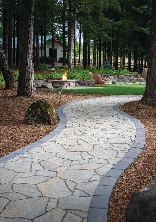 Curving stone garden path through tall trees, bordered by mulch and grass, with a small house in the background