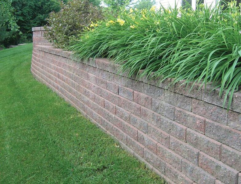 A slightly curved retaining wall made of stacked, textured reddish-brown stones, bordered by a green lawn and plants.