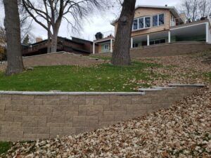 A beige house sits atop a landscaped yard with multiple tiers of stone retaining walls and mature trees.