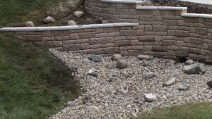A multi-tiered retaining wall made of tan stone blocks with a gravel drainage area in the foreground and a grassy hill.