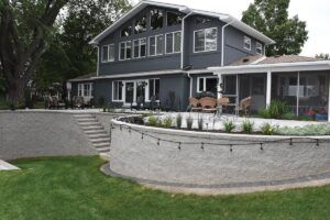 A two-story dark gray house with a stone retaining wall, patio, and stairs leading to a landscaped backyard.