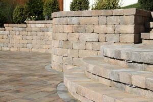 Tan curved stone retaining wall steps leading to a matching stone patio with green bushes in the background.