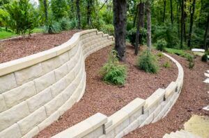 A curved, multi-tiered beige stone retaining wall holding back terraced garden beds filled with mulch and small shrubs.