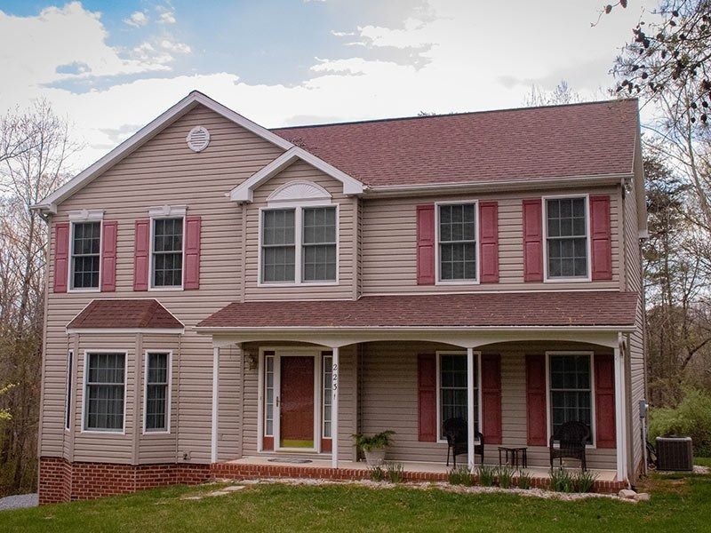 Two-story beige house with red shutters and roof, porch with black chairs, set in a green yard.