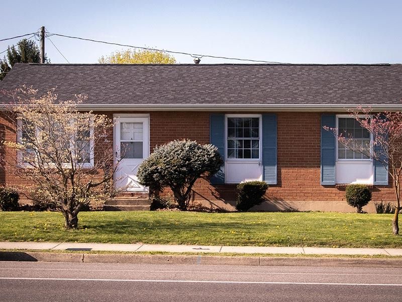 A brick home with blue shutters and white trim, trees and bushes line the front lawn.