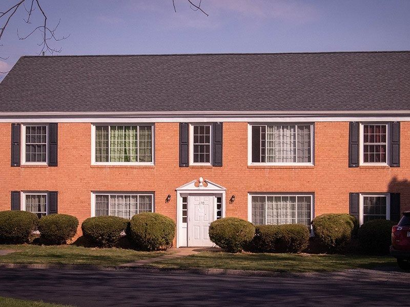 Red brick apartment building with black shutters, gray roof, and manicured bushes.